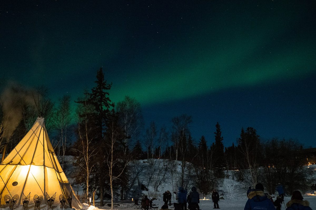 Green aurora spreading over a lit teepee tent and conifers on a winter night in Yellowknife